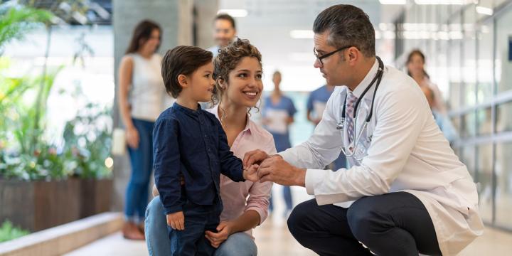Male pediatrician talking to child patient who is standing next to his mom, all are smiling