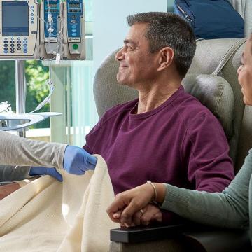 Nurse pulling a blanket up for a man in a hospital chair. A woman holds his hand.