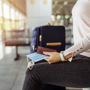 image from neck to knee of a woman sitting on a bench with a rolling suitcase in the background and passports in hand