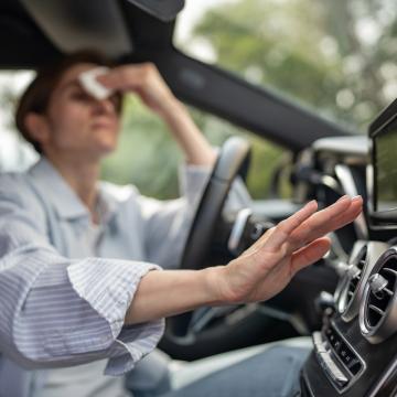 image of a woman in a car, wiping her forehead with a cloth and checking the air conditioning vents with her other hand