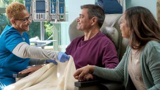 Nurse pulling a blanket up for a man in a hospital chair. A woman holds his hand.