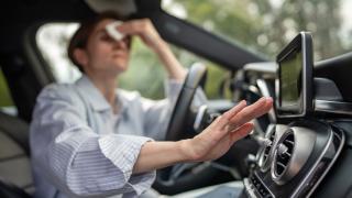 image of a woman in a car, wiping her forehead with a cloth and checking the air conditioning vents with her other hand