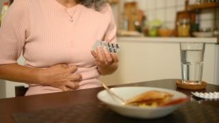 woman sitting at table holding medication and clutching stomach