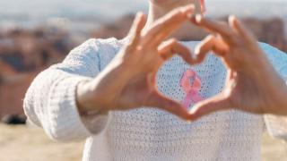 woman making a heart symbol with hands in front of pink ribbon on sweater